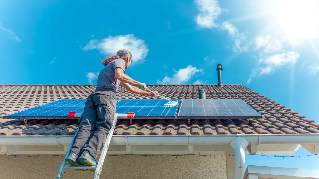 A technician using tools to clean solar panels on a pitched roof with safety equipment and ropes.