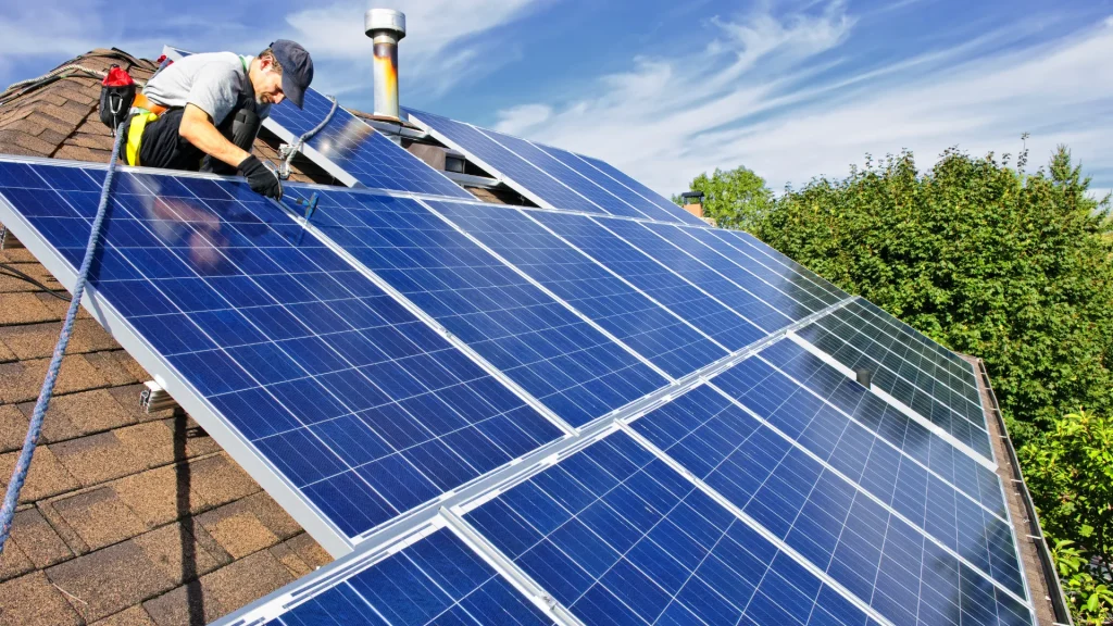 A worker on a ladder cleaning solar panels on a pitched roof under a sunny sky.