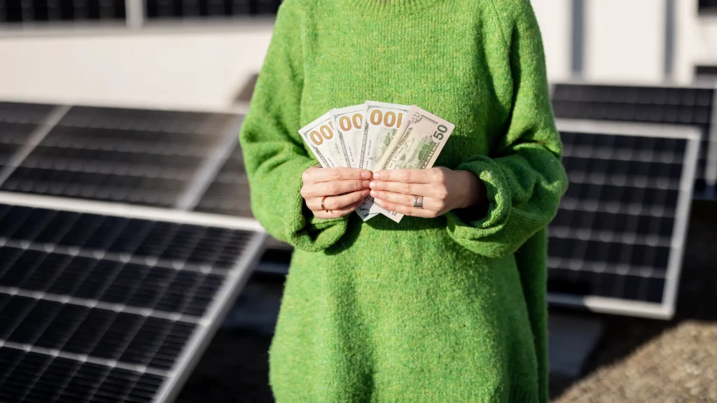 Person holding dollar bills in front of solar panels, symbolizing cost savings from Trina Solar panel energy generation.